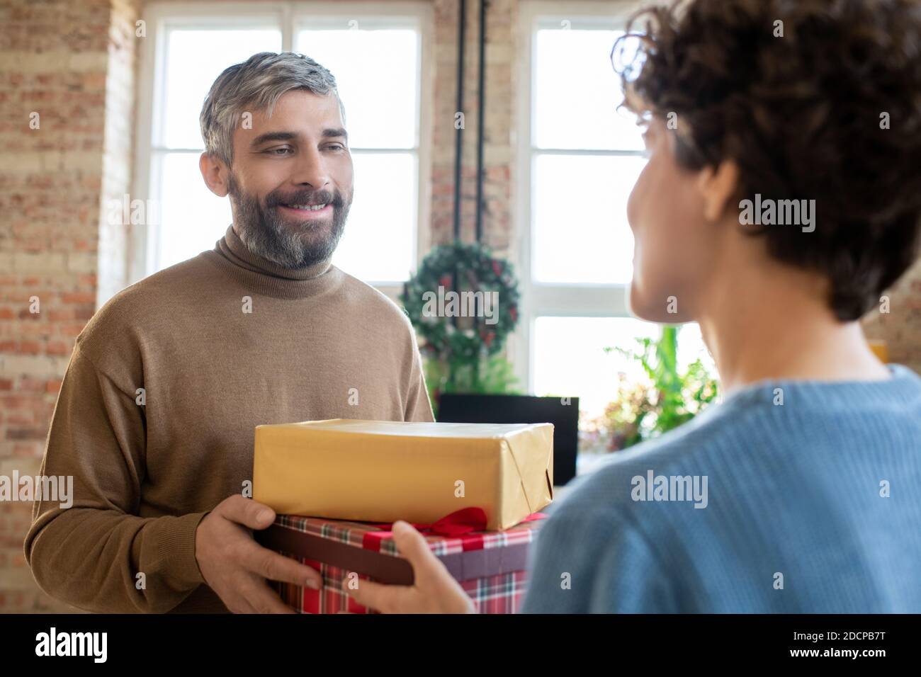 Glücklicher bärtiger Mann, der während seiner Zeit zwei Geschenke an seine Frau übergibt Sie mit einem Lächeln anblickend Stockfoto