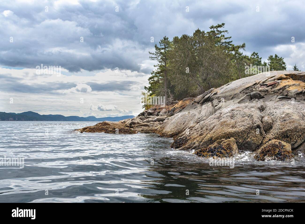An einem unruhigen Tag ragt ein felsiger Punkt in das gekräuselte Wasser, während drohende Wolken über den Inseln in der Ferne zu schweben scheinen. Stockfoto