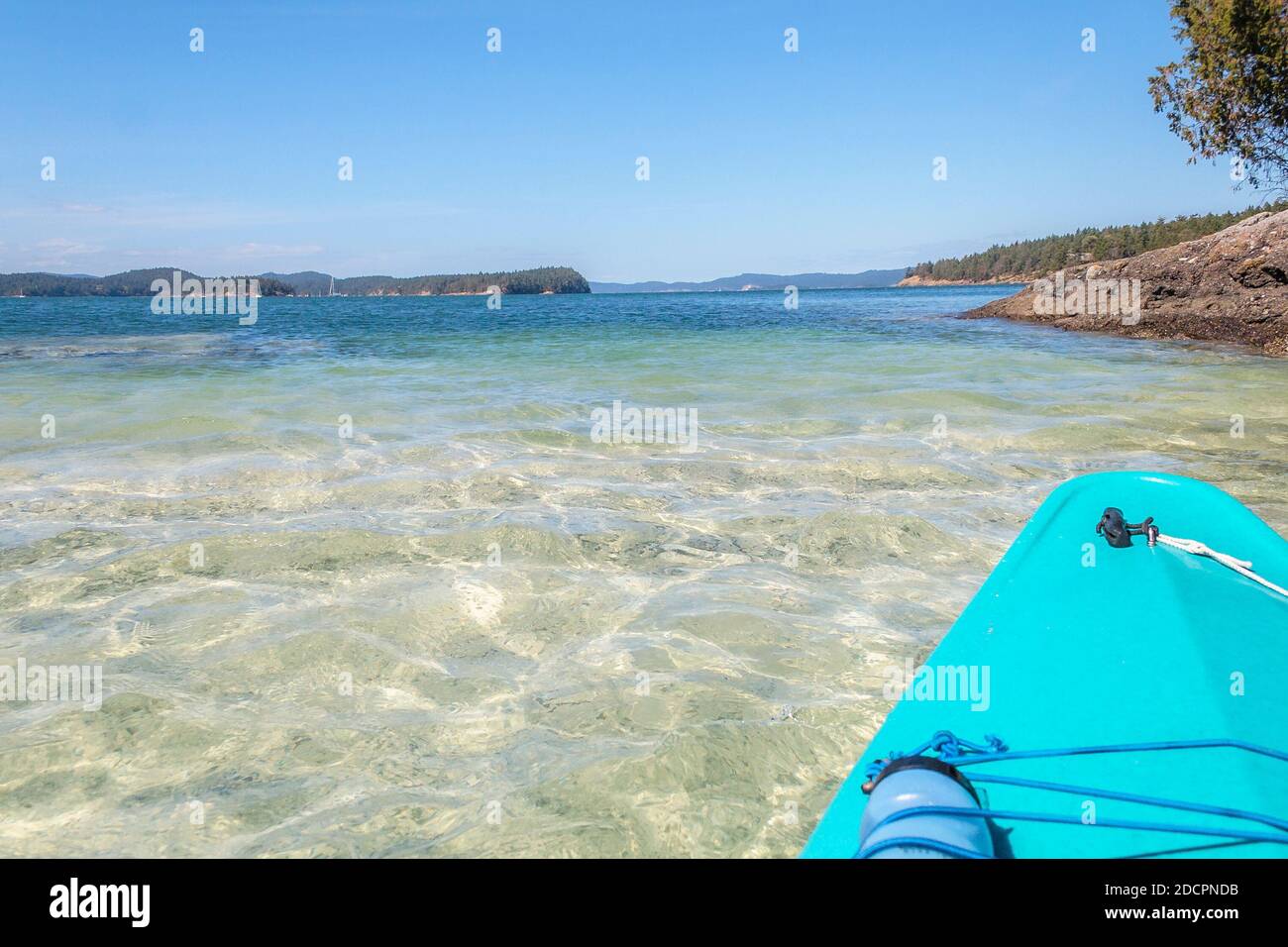 Ein Schlamm und Muschelboden wird durch das klare Wasser gesehen, wie ein Kajak über eine Untiefe bewegt, mit tiefblauem Wasser und Inseln dahinter (British Columbia). Stockfoto
