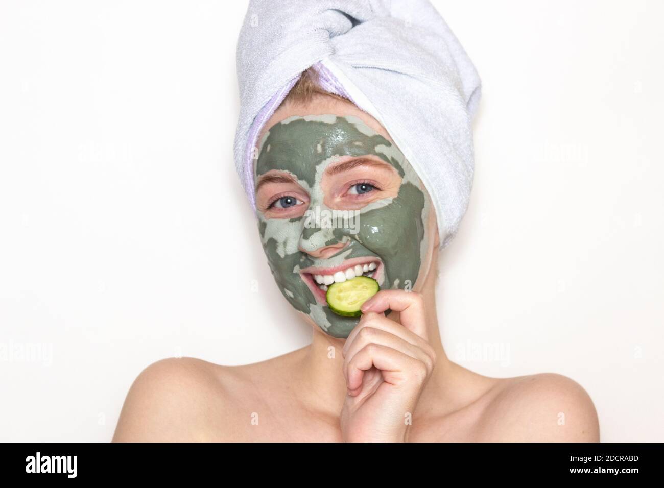 Eine junge Frau mit einem Handtuch auf dem Kopf und einer Tonmaske auf ihrem Gesicht und einem Stück Gurke. Hautpflege zu Hause. Weißer Hintergrund. Stockfoto