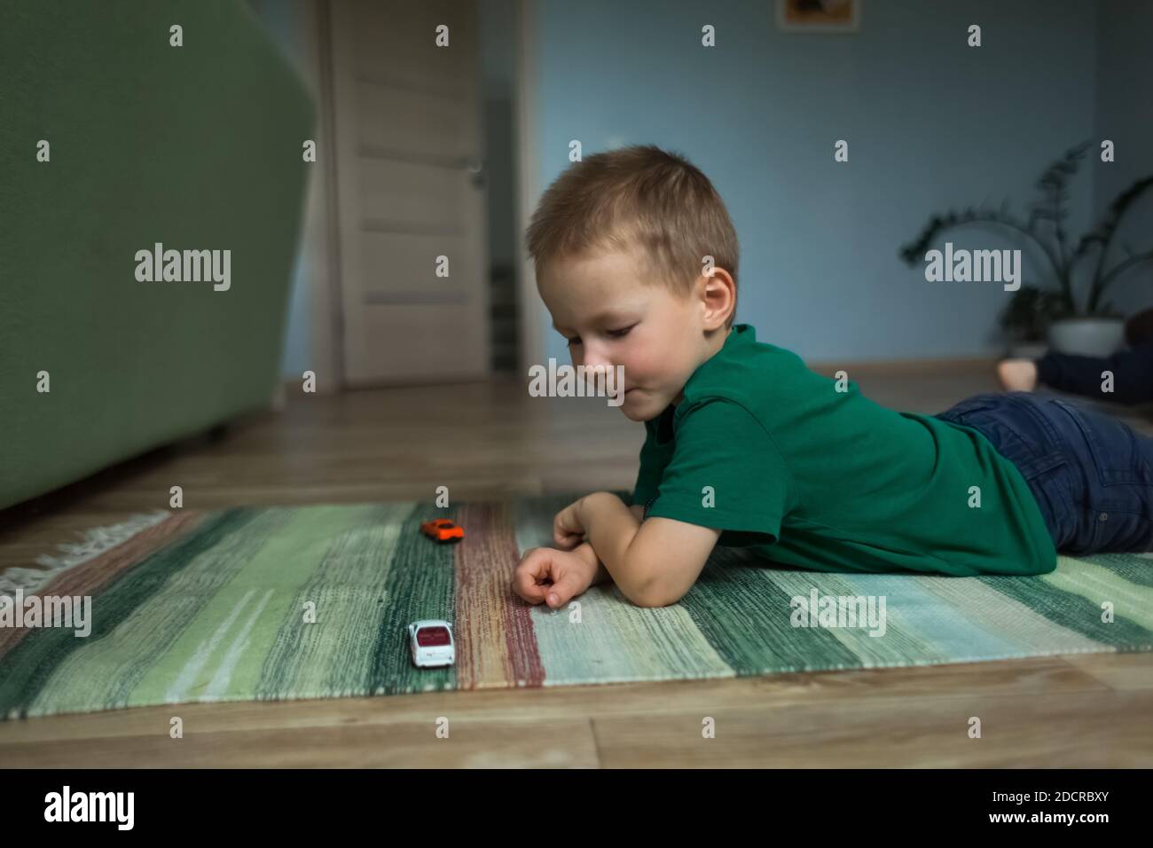 Hübscher Junge, der auf dem Boden auf dem Teppich liegt, spielt mit Spielzeugautos. Stockfoto