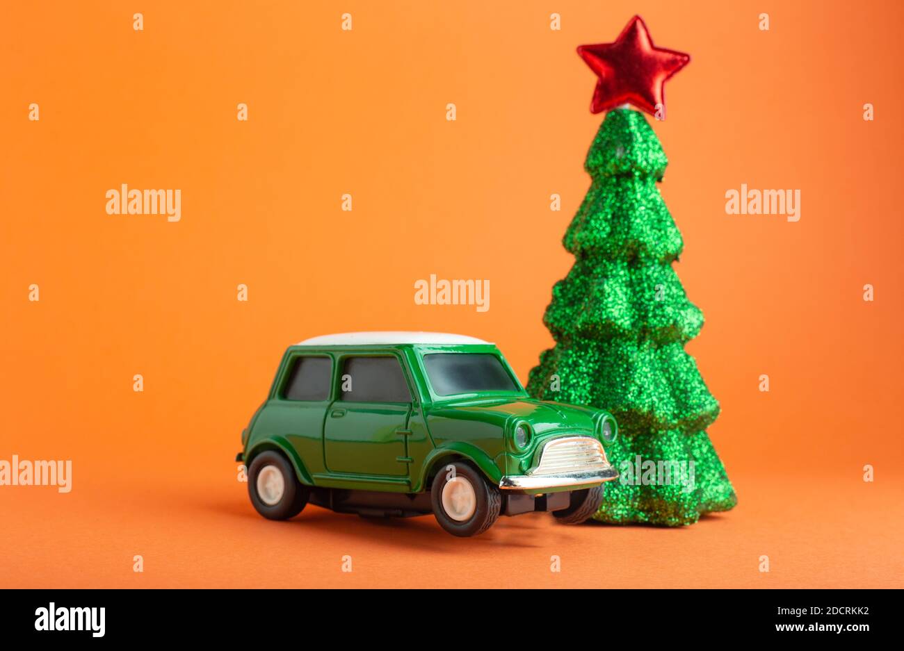 Weihnachtsbaum mit rotem Stern auf der Oberseite in der Nähe von grünen Auto Spielzeug. Orangefarbener Hintergrund. Kreative Miniatur Weihnachtsbaum und Auto. Konzept für die Zustellung von Geschenken Stockfoto