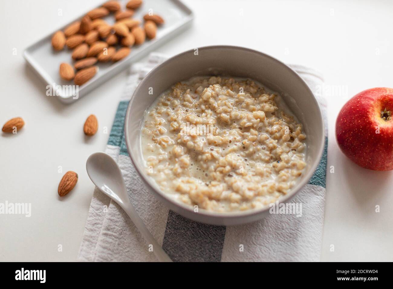 Hausgemachter Haferbrei Haferbrei mit Nussmandeln in einer Schüssel, Keramiklöffel, roter Apfel auf weißem Hintergrund. Health Frühstück Essen Konzept Stockfoto