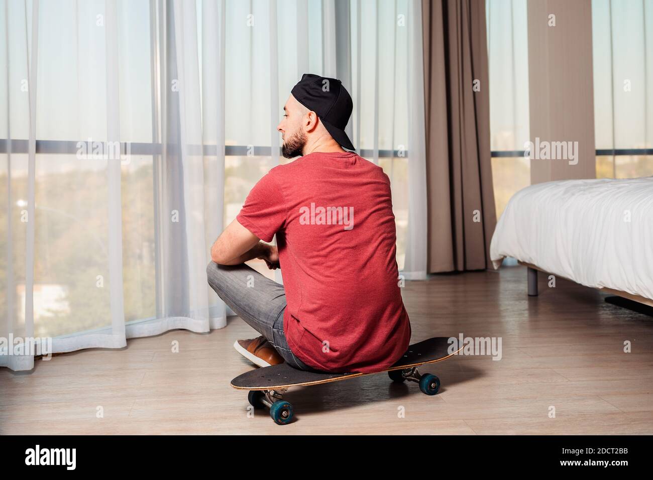 Ein junger Mann sitzt auf einem Longboard und schaut aus dem Fenster. Inneneinrichtung. Rückansicht. Das Konzept von Quarantäne und Aktivität zu Hause. Stockfoto
