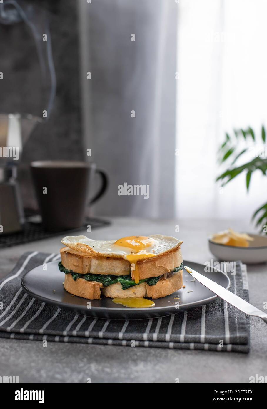 Ein Sandwich mit einem Eigelb, das aus einem Spiegelei fließt. Leckeres Frühstückssandwich mit Spinat und Kaffeemaschine mit heißem Kaffee. Stockfoto