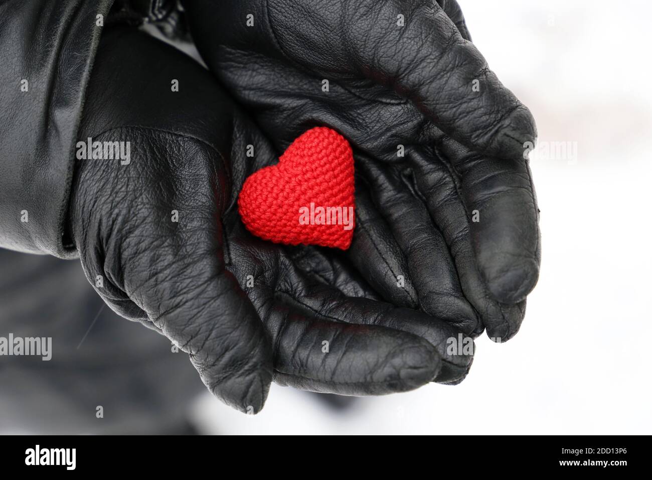 Rot gestricktes Herz in weiblichen Händen in schwarzen Lederhandschuhen auf Schneehintergrund. Konzept der romantischen Liebe, Valentinstag, Gesundheitsversorgung im Winter Stockfoto