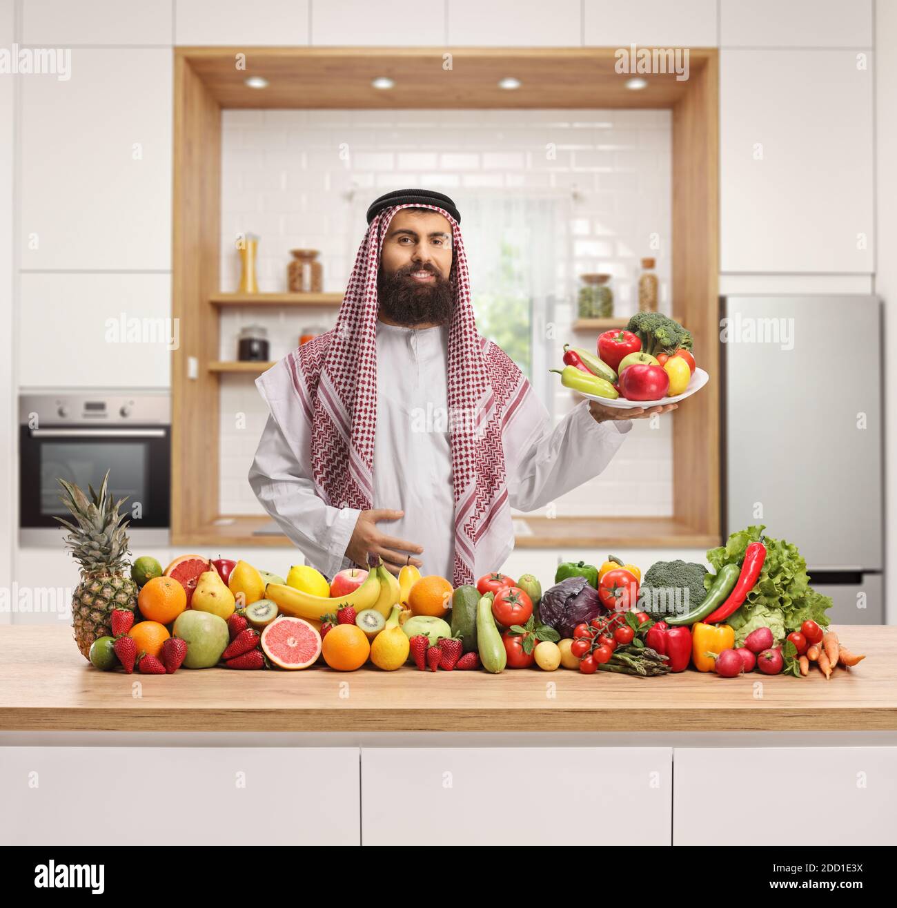 Traditioneller saudi-arabischer Mann in einer Küche, der einen Teller hält Von Obst und Gemüse und stehen hinter einem Haufen von Obst und Gemüse Stockfoto
