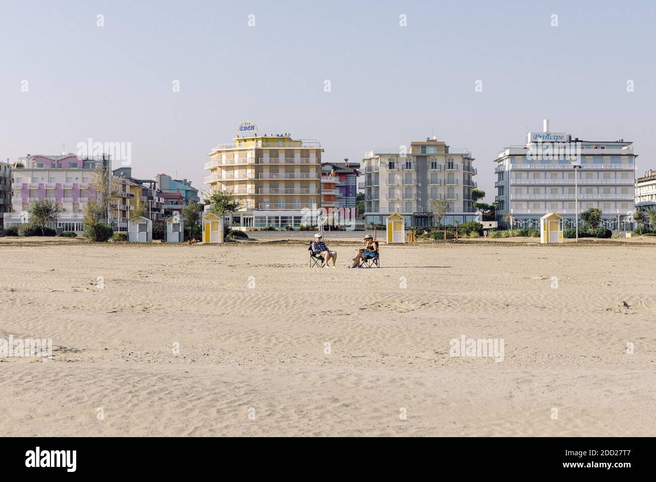 Zwei alte Leute, die außerhalb der Saison auf einem Liegestuhl an einem Wüstenstrand sitzen, mit hohen Gebäuden und Hotels im Hintergrund Stockfoto