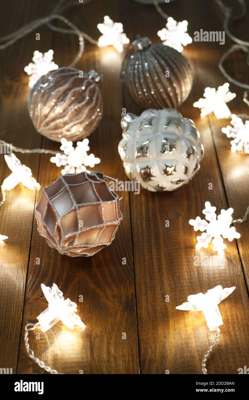 Rosa, graue und weiße Weihnachtskugeln liegen auf einem braunen Holztisch zusammen mit einer leuchtenden Girlande. Nahaufnahme Stockfoto
