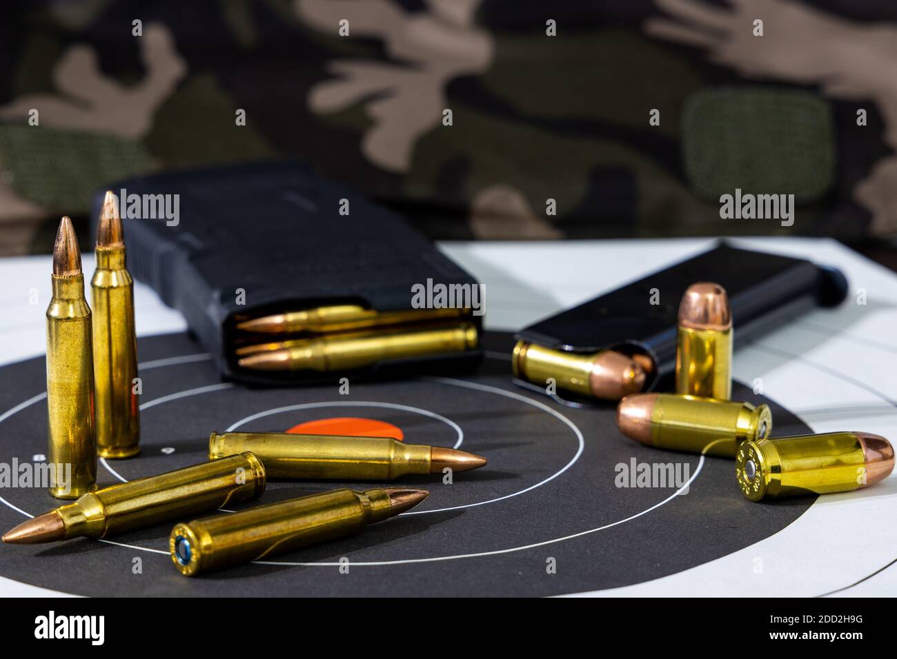 Geladene Gewehr- und Pistolenmagazine mit losen Munition, die auf einem Papierziel mit Tarnhintergrund verstreut sind. Stockfoto