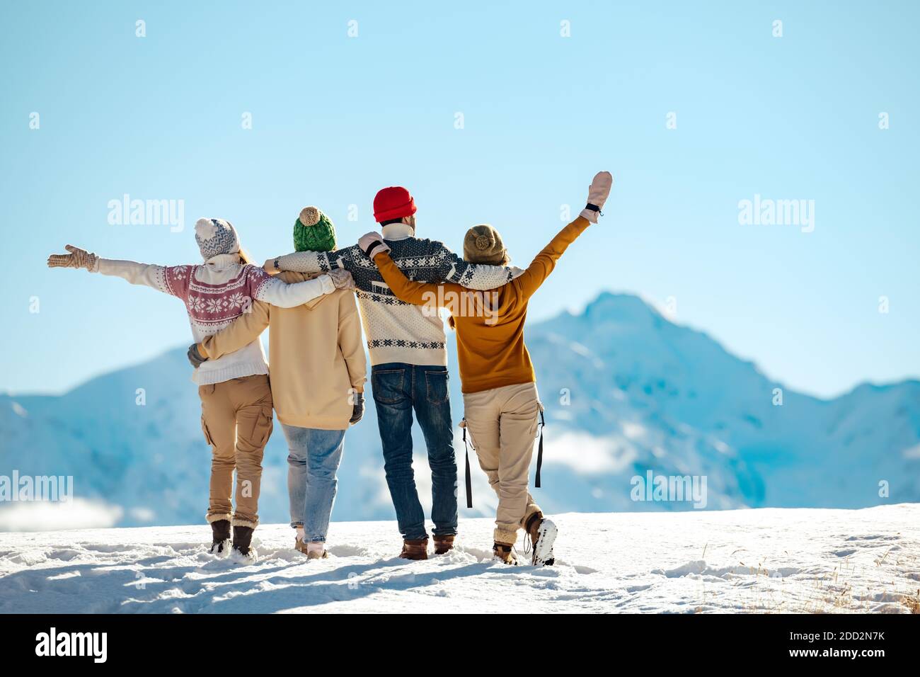 Vier glückliche Freunde stehen und umarmen sich an sonnigen Tagen gegen schneebedeckte Berge. Winterurlaub Konzept Stockfoto