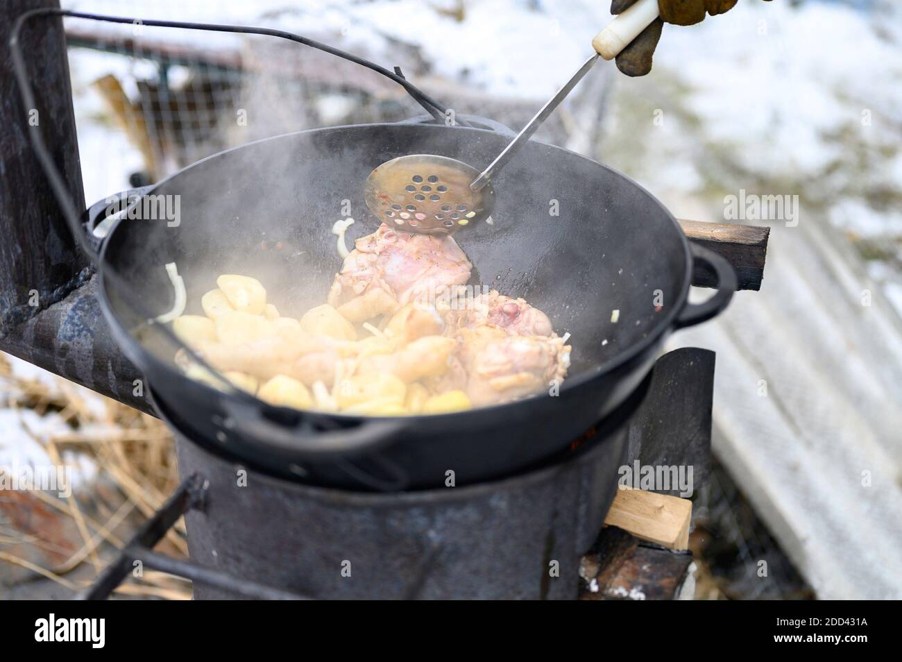 Kochen im Winter in einem Kessel im Freien auf einem Feuer auf einem gusseisernen Ofen. Essen aus Kartoffeln, Zwiebeln und Hühnerfleisch wird in einem Gusseisen zubereitet Stockfoto