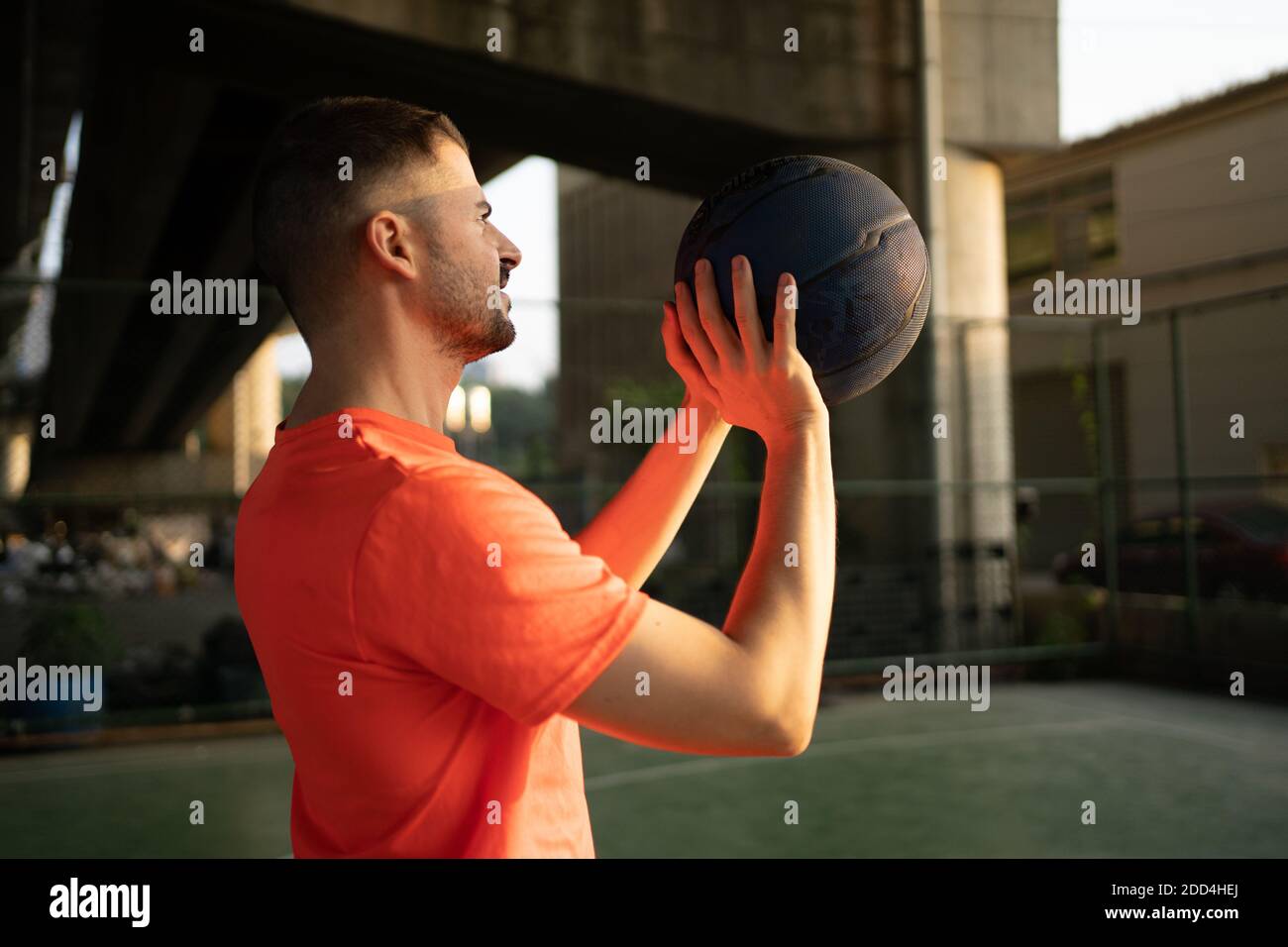 Kaukasischer junger Mann spielt Basketball allein auf städtischen Strecke unter highway Bridge in orangefarbenem T-Shirt Stockfoto