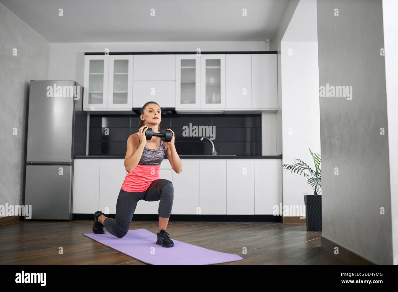 Fit Mädchen tragen Sportkleidung Training zu Hause am Morgen mit Hantel. Konzept der Home-Workout, Gewichtsverlust. Seitenansicht einer starken kaukasischen jungen Frau, die in der Küche Ausfallschritte auf einer Matte praktiziert. Stockfoto