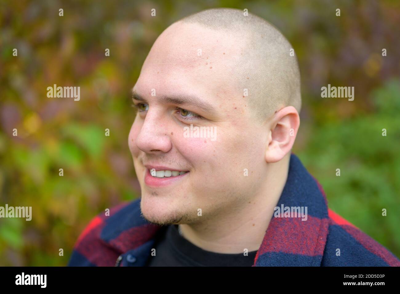 Junger Mann mit rasierten Kopf, der mit einer Ruhe zur Seite schaut Durchdachtes Lächeln im Freien gegen Grün Stockfoto