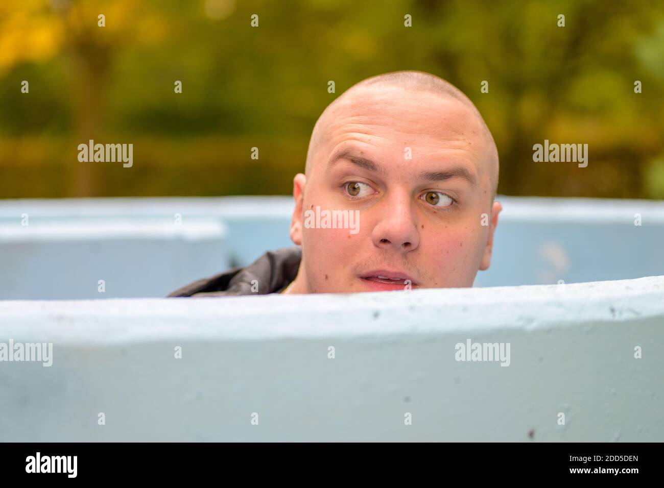 Junger Mann mit rasierten Kopf Blick über den Rand des Ein Outdoor-Tank in einem Park mit faszinierten Ausdruck und Erhöhte Augenbrauen in Nahaufnahme Stockfoto