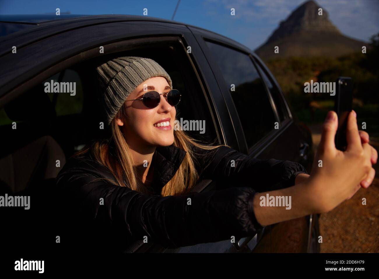 Junge Frau trägt Sonnenbrille und Mütze Hut auf der Reise Urlaub Foto aus dem Mietwagen Fenster auf dem Handy Telefon Stockfoto