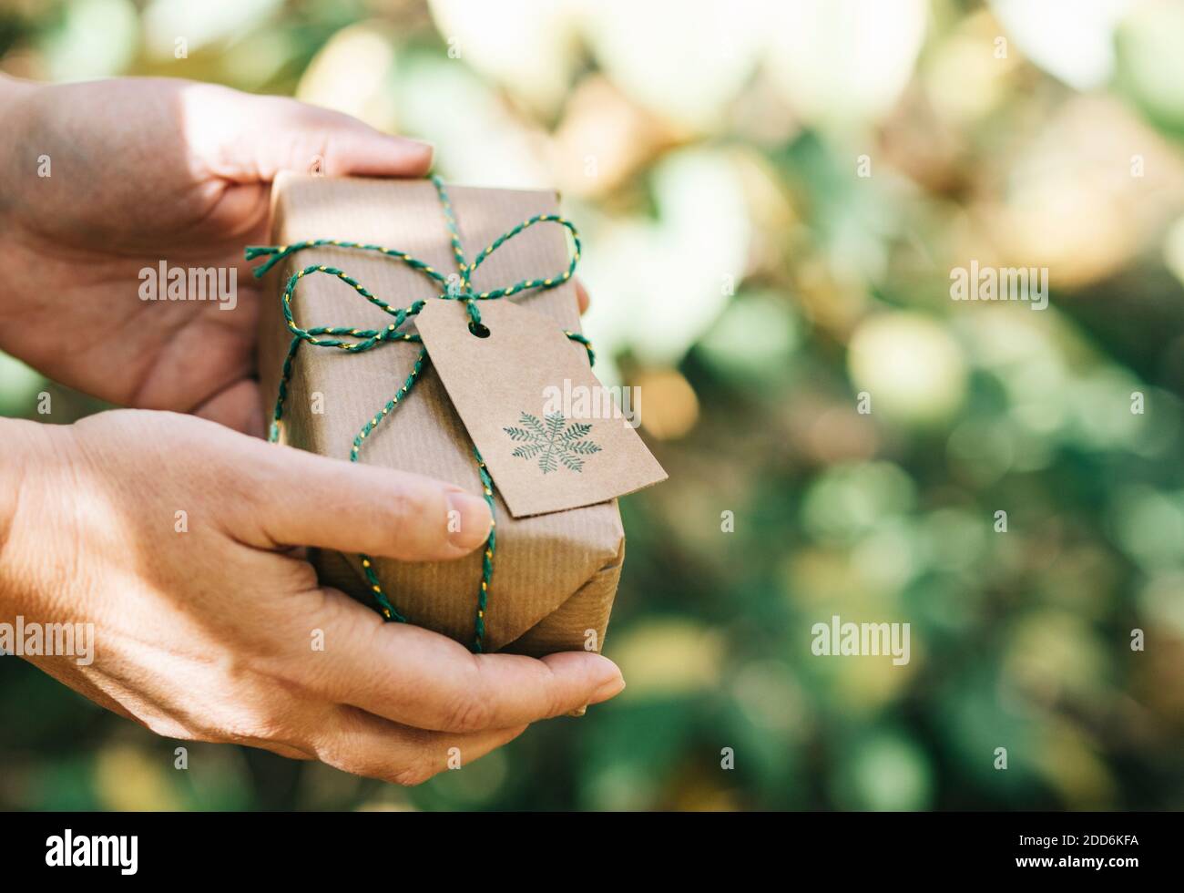 Hände liefern ein Weihnachtsgeschenk verpackt in recyceltem Papier mit der Natur verschwommen. Ökologisches und handgefertigtes Geschenkkonzept. Stockfoto