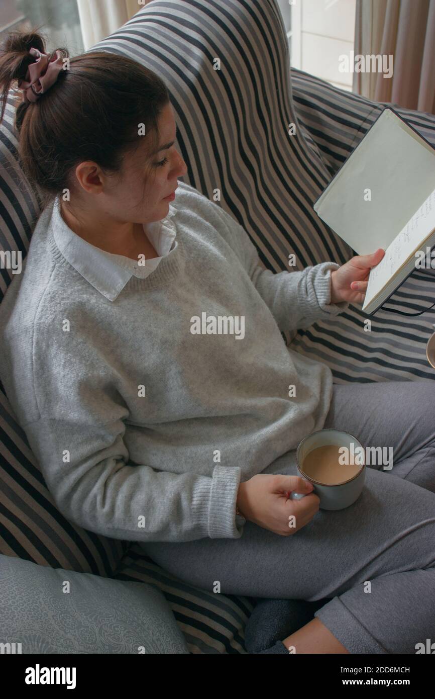 Hispanische Brünette Frau genießt ihr heißes Getränk auf der Couch sitzend, während sie ein Buch in einem warmen Outfit an einem Wintertag liest. Winterfotografie 2020. Stockfoto