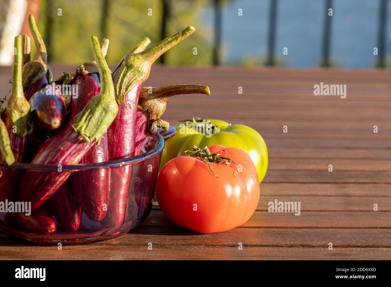 Konzentrieren Sie sich auf das Sonnenlicht, das auf eine rote Tomate reflektiert. Auf dem hölzernen Tisch eine Gruppe von kleinen Auberginen im Glas und eine andere grüne Frucht. Hintergrund für das Essen Stockfoto