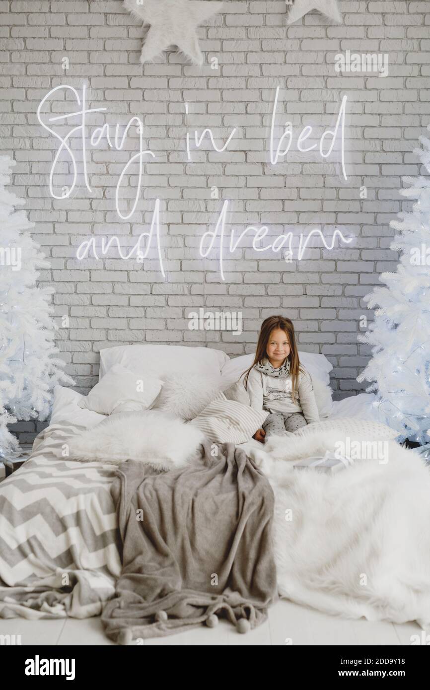 Nettes kleines Mädchen sitzt auf dem Bett zu Hause mit weißen Weihnachtsbäumen auf dem Hintergrund. Stockfoto