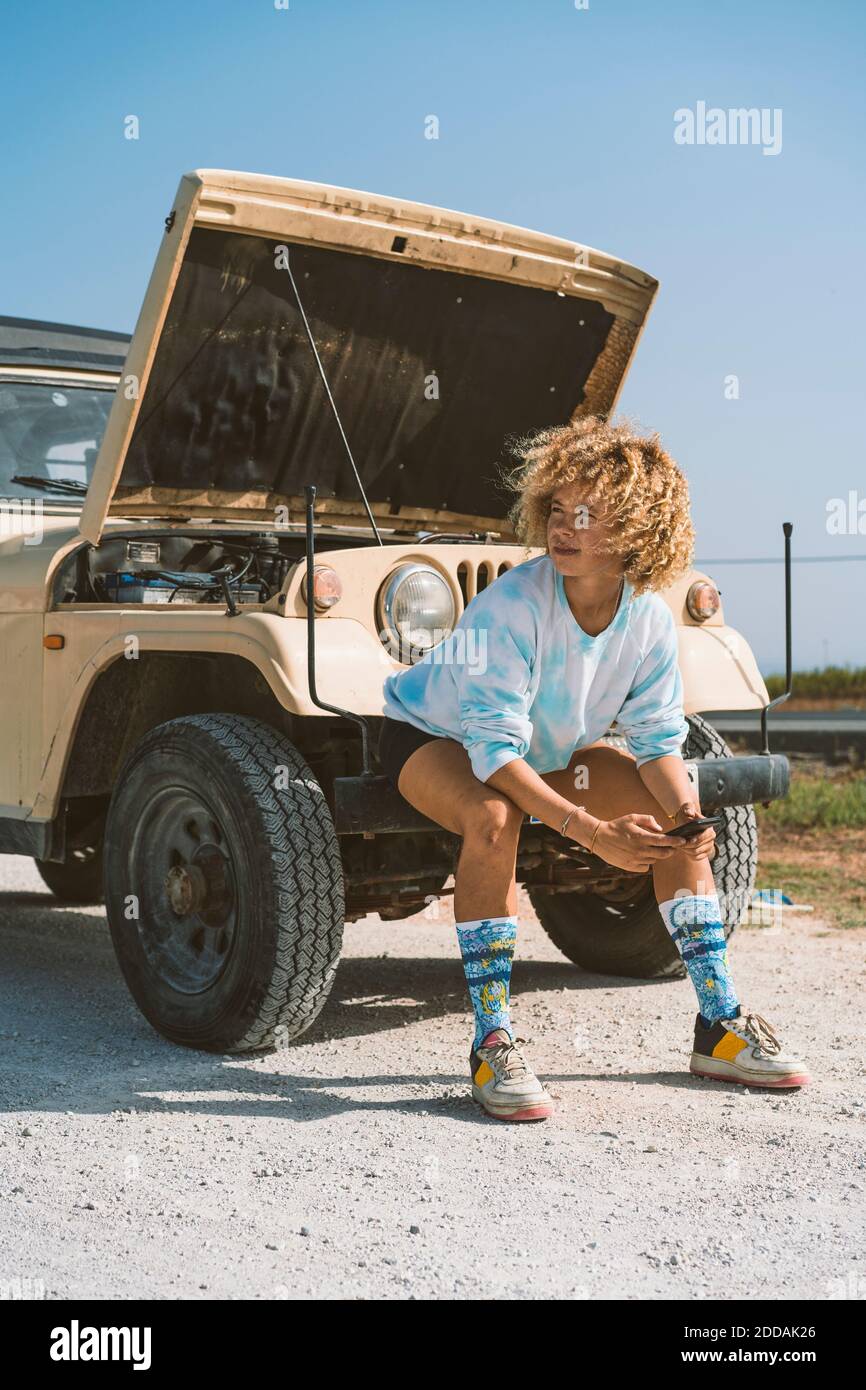 Junge blonde Afro-Frau schaut weg, während sie auf der Stoßstange sitzt Des abgebrochenen Fahrzeugs an sonnigen Tagen Stockfoto