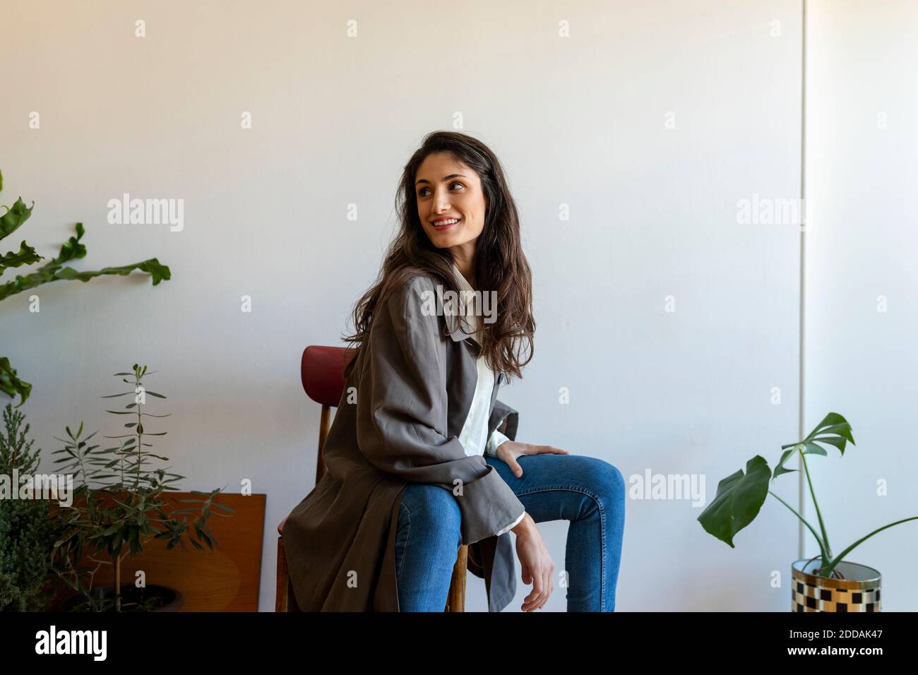 Lächelndes weibliches Modell, das wegschaut, während es auf Stuhl sitzt Zu Hause Stockfoto