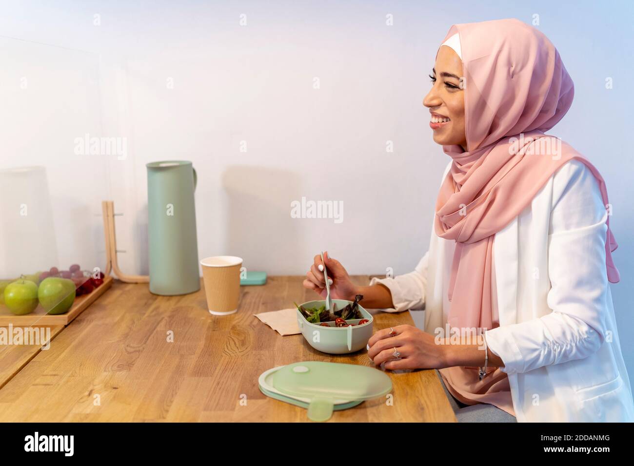 Lächelnde junge Frau, die Essen isst, während sie an der Bildschirmwand sitzt Tisch im Büro Stockfoto