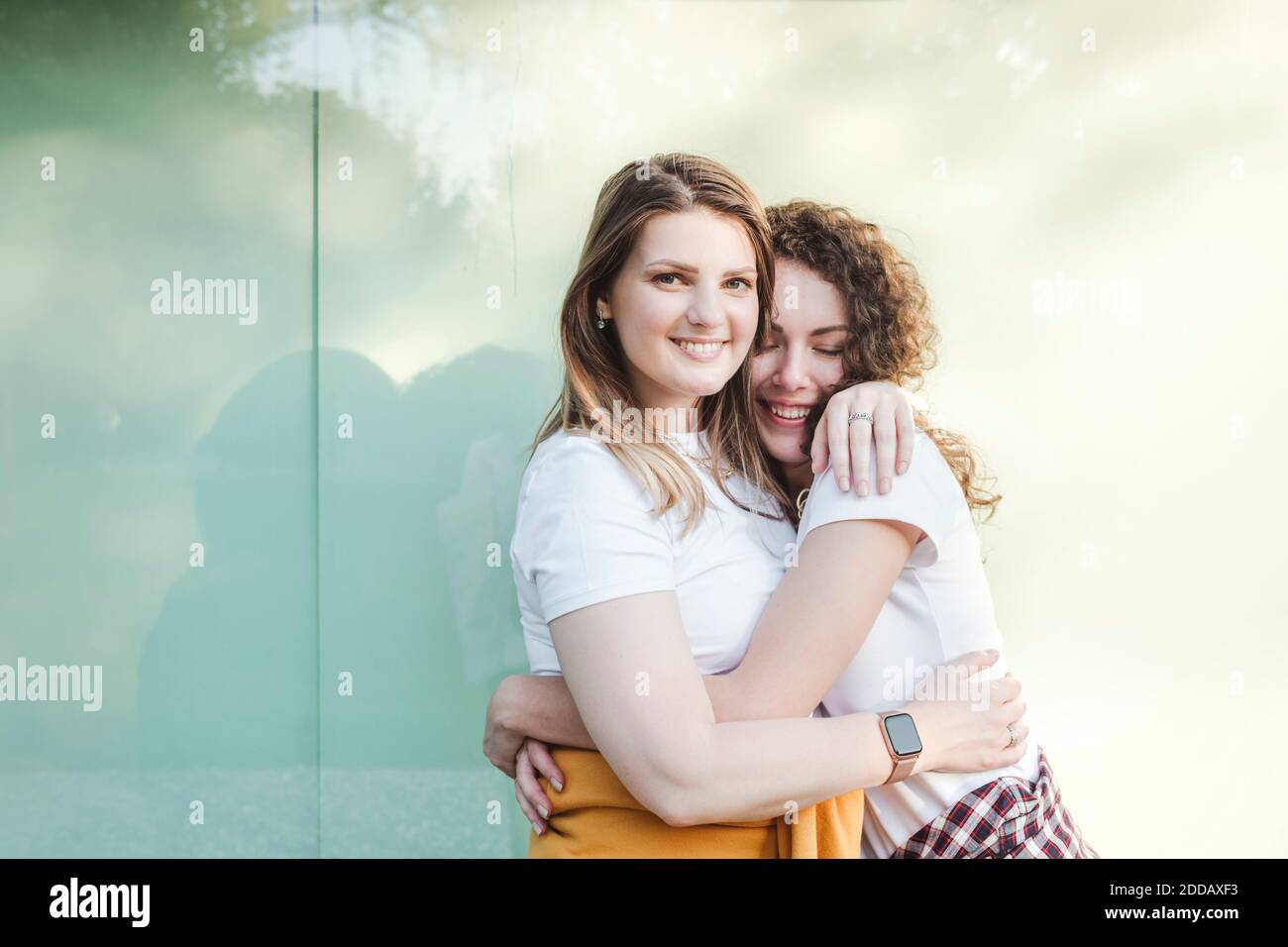 Lächelnd schöne Frau wird von jungen weiblichen Freund gegen umarmt Wand Stockfoto