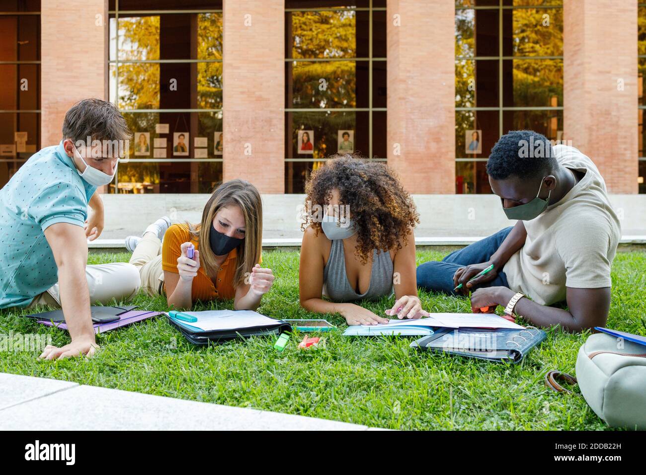 Männliche und weibliche Freunde tragen Sicherheitsmaske während des Studiums über Gras auf dem Universitätscampus Stockfoto