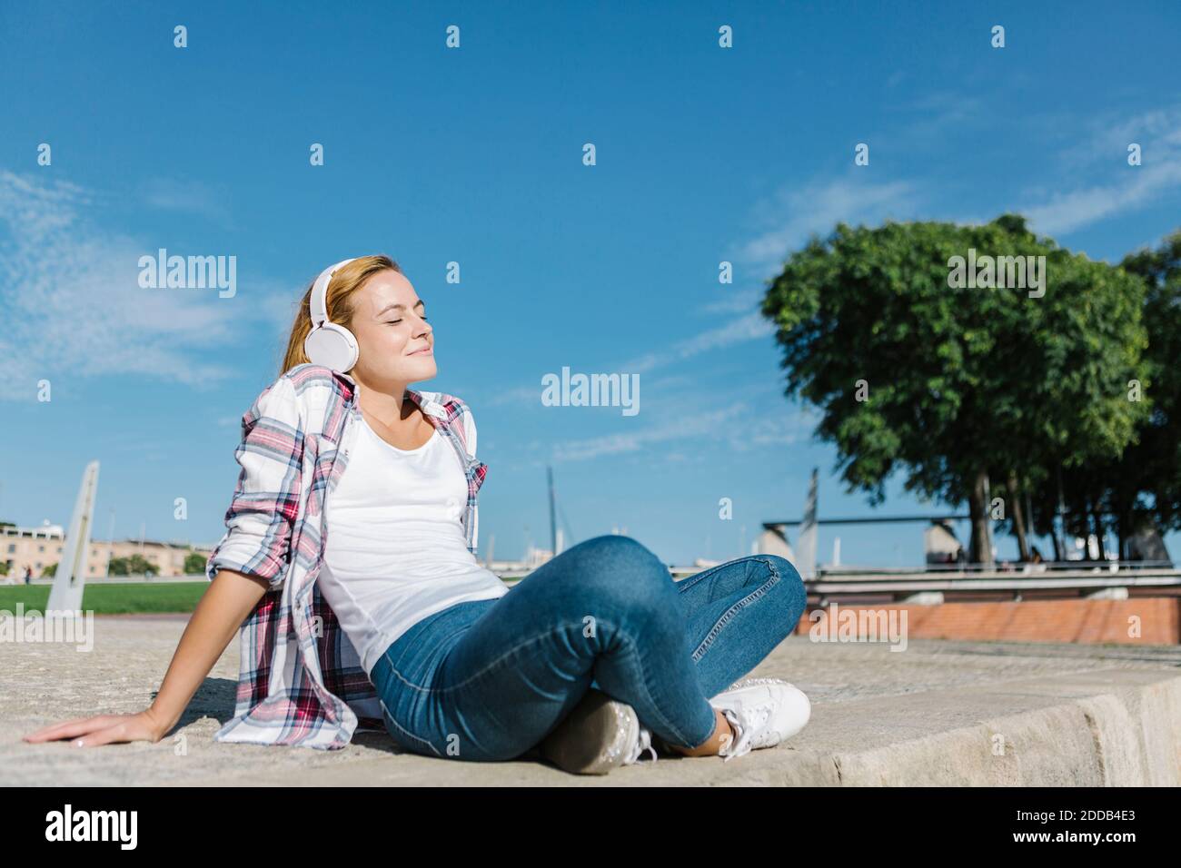 Frau, die Musik hört, während sie an sonnigen Tagen auf dem Fußweg sitzt Stockfoto