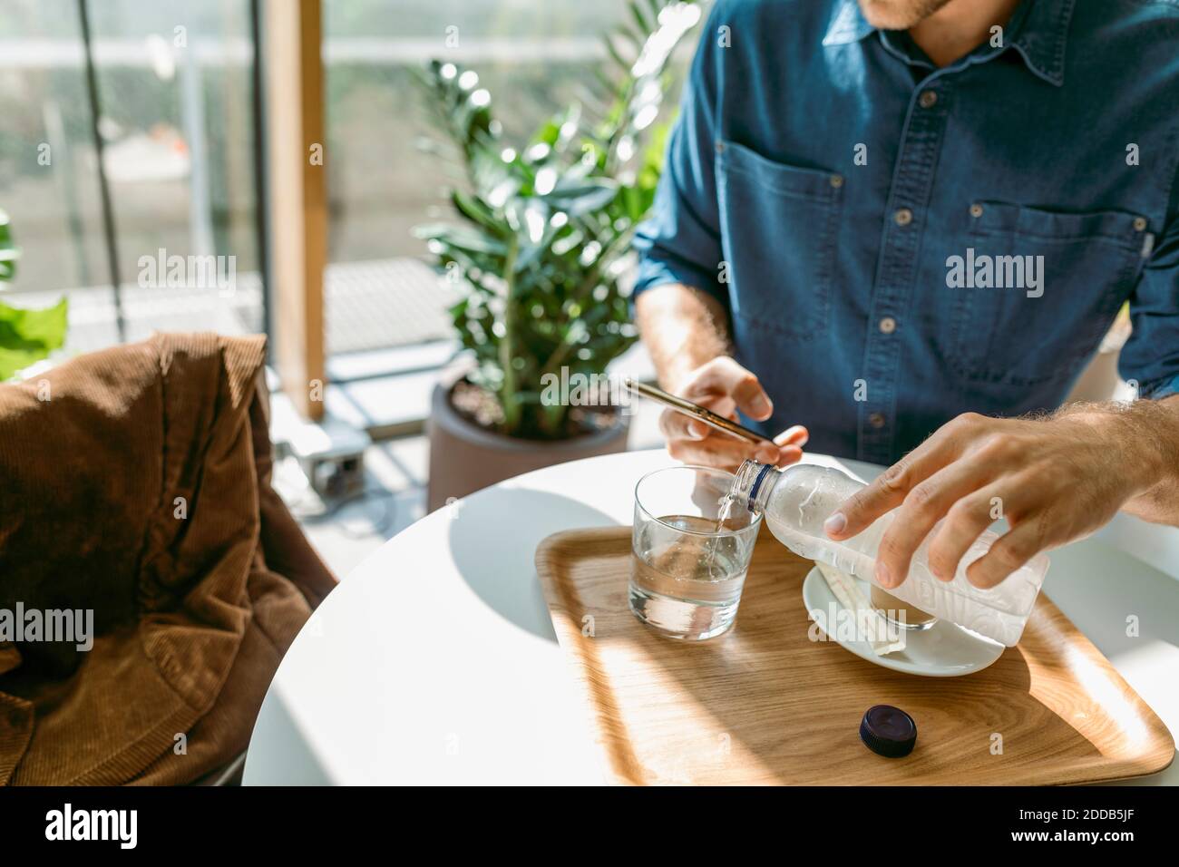 Junger Geschäftsmann mit Smartphone, während Wasser in Glas gießen Am Tisch im Café Stockfoto