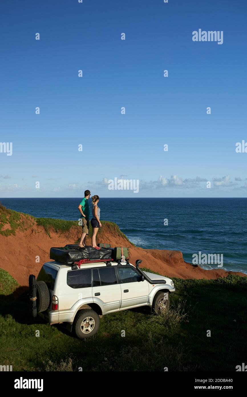 Mittelerwachsenes Paar, das auf dem 4x4-Dach steht, während es sich anschaut Meer gegen blauen Himmel Stockfoto