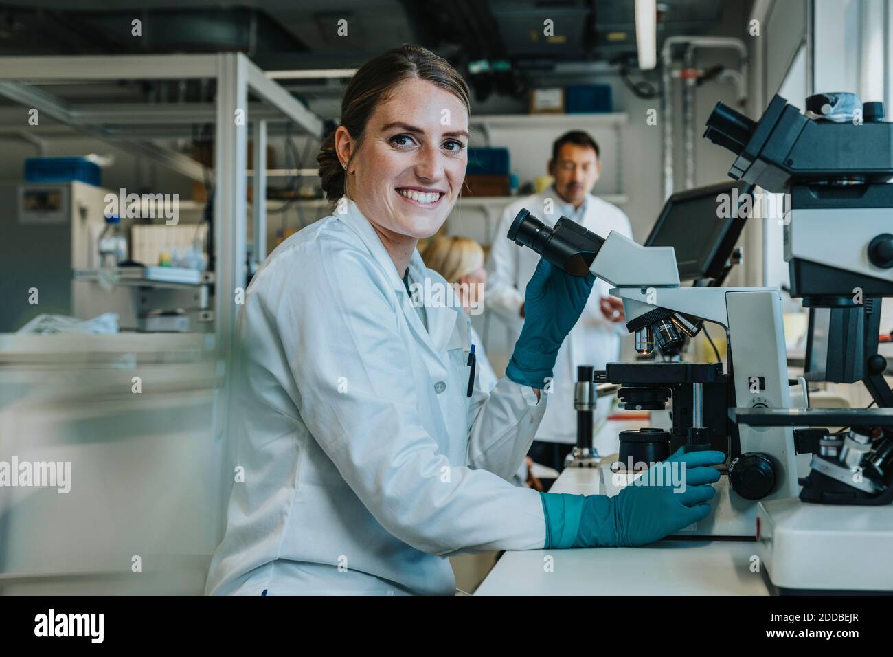 Lächelnde Frau, die menschliches Gehirn analysiert, gleiten unter das Mikroskop, während sie sitzt Mit Wissenschaftlern im Hintergrund im Labor Stockfoto