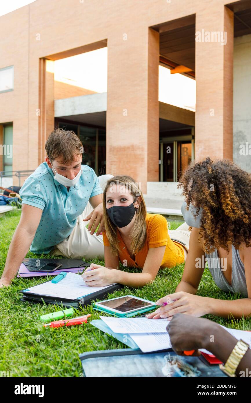 Studentin mit Freunden, die eine Sicherheitsmaske tragen, während sie über das Studium lernt Gras auf dem Universitätscampus Stockfoto