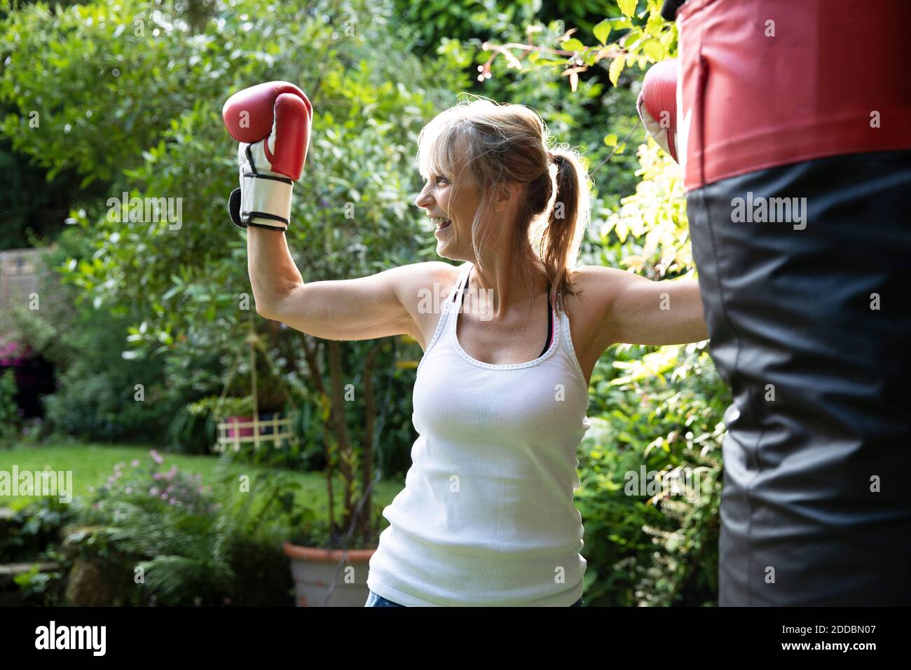 Ältere Frau lächelt, während sie die Muskeln im Boxhandschuh im Stehen anbiegt Im Hinterhof am sonnigen Tag Stockfoto