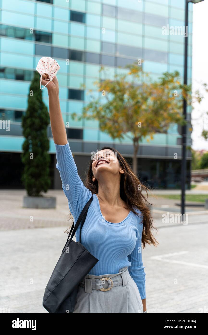 Glückliche Frau mit Geldbörse hält schützende Gesichtsmaske in der Luft Gegen Gebäude während COVID-19 Stockfoto