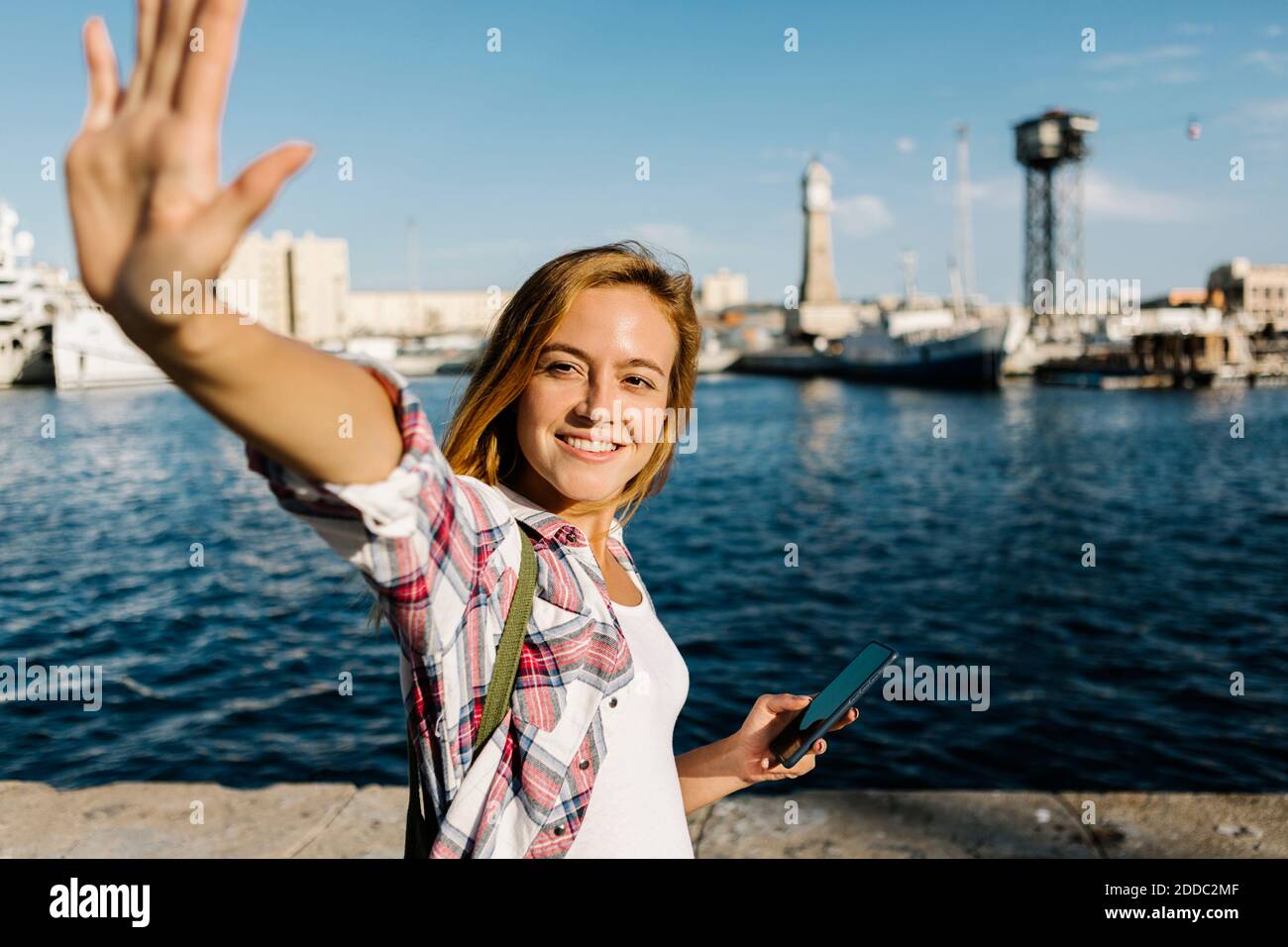 Frau winkt, während sie das Smartphone am Meer hält Sonniger Tag Stockfoto