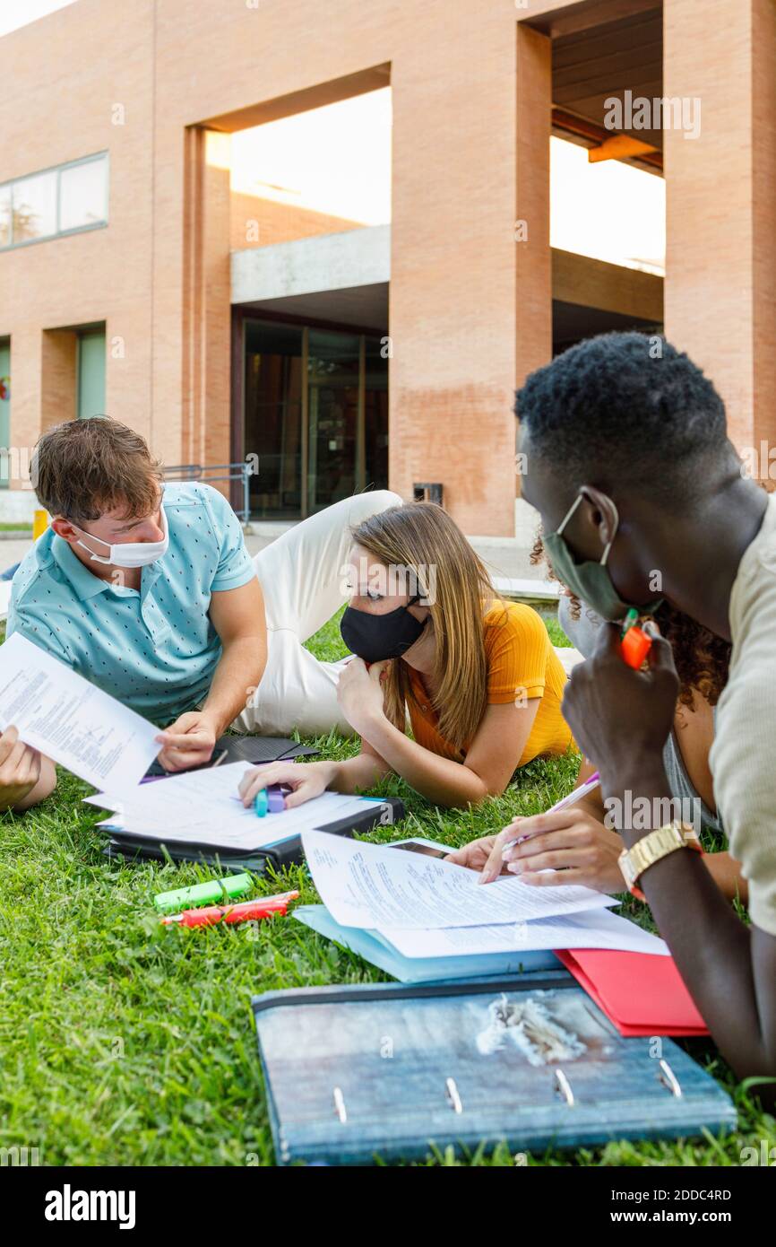 Studentin mit Freunden, die eine Sicherheitsmaske tragen, während sie über das Studium lernt Gras auf dem Universitätscampus Stockfoto