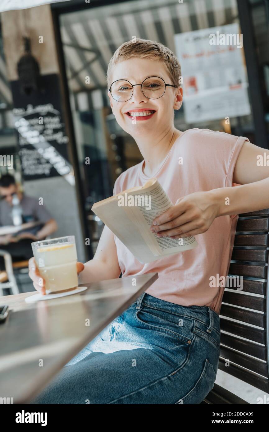 Lächelnde Frau mittleren Erwachsenen Buch lesen, während auf dem Bürgersteig sitzen Mit Kaffee trinken an sonnigen Tag Stockfoto