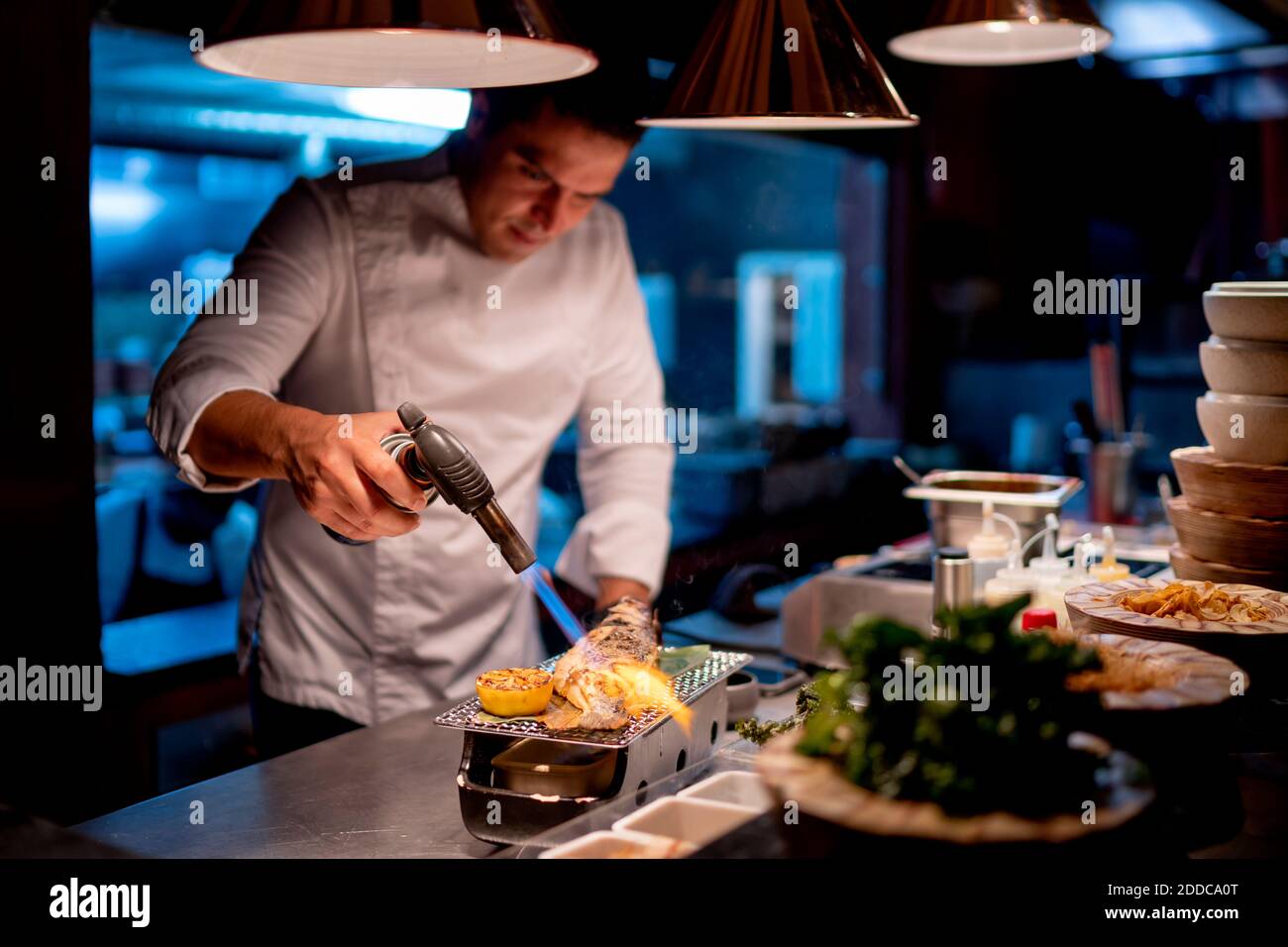 Männlicher Koch mit Blasfackel während der Zubereitung von Meeresfrüchten in der Küche Im Restaurant Stockfoto