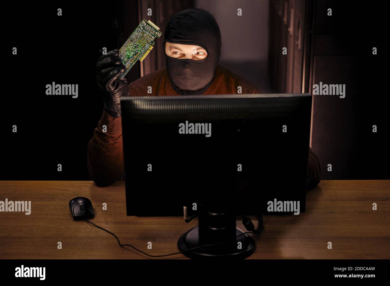 Mann in schwarzer Maske mit einer Leiterplatte von einem Hacker-Gerät in der Hand sitzt an einem Computer. Konzept von Datensicherheitsproblemen und Netzwerk-Hacking Stockfoto