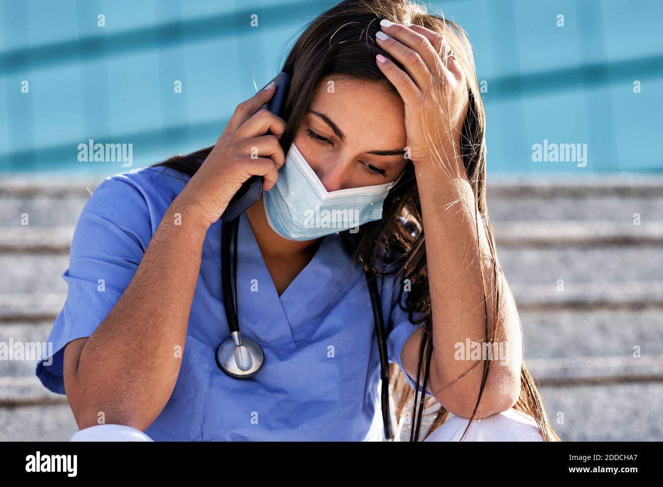 Junge Arzt trägt Gesichtsmaske reden auf Handy, während Sitzen auf der Treppe gegen das Krankenhaus Stockfoto