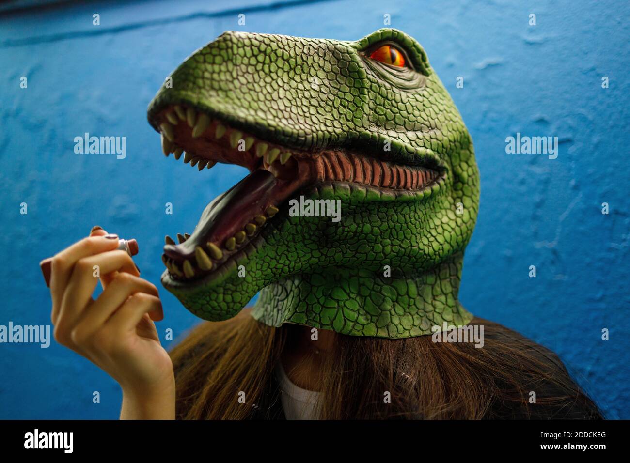 Frau, die Lippenstift trägt, während sie Dinosauriermaske an der blauen Wand trägt Stockfoto
