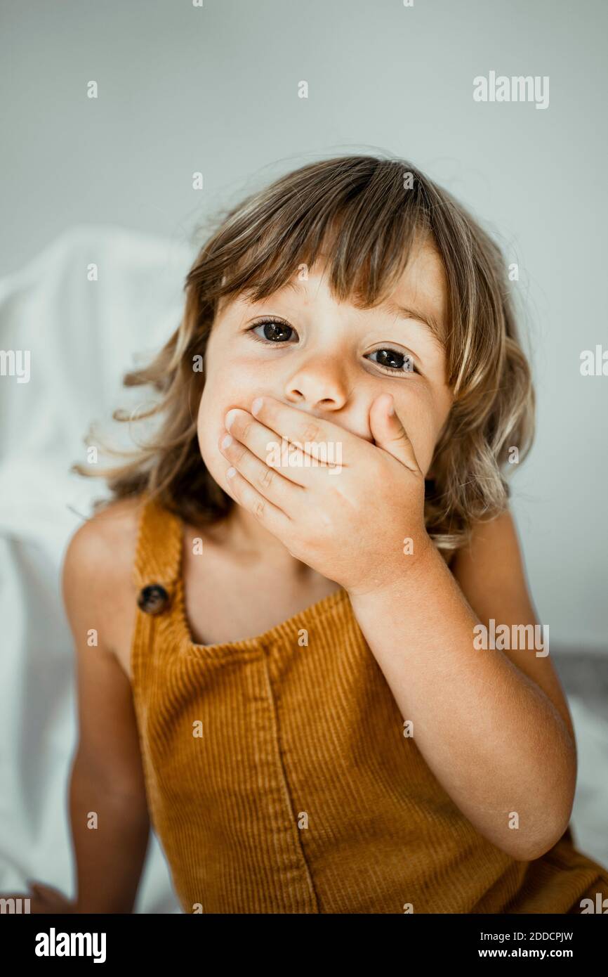 Nettes Mädchen, das den Mund mit den Händen bedeckt, während es an der Wand sitzt Zu Hause Stockfoto