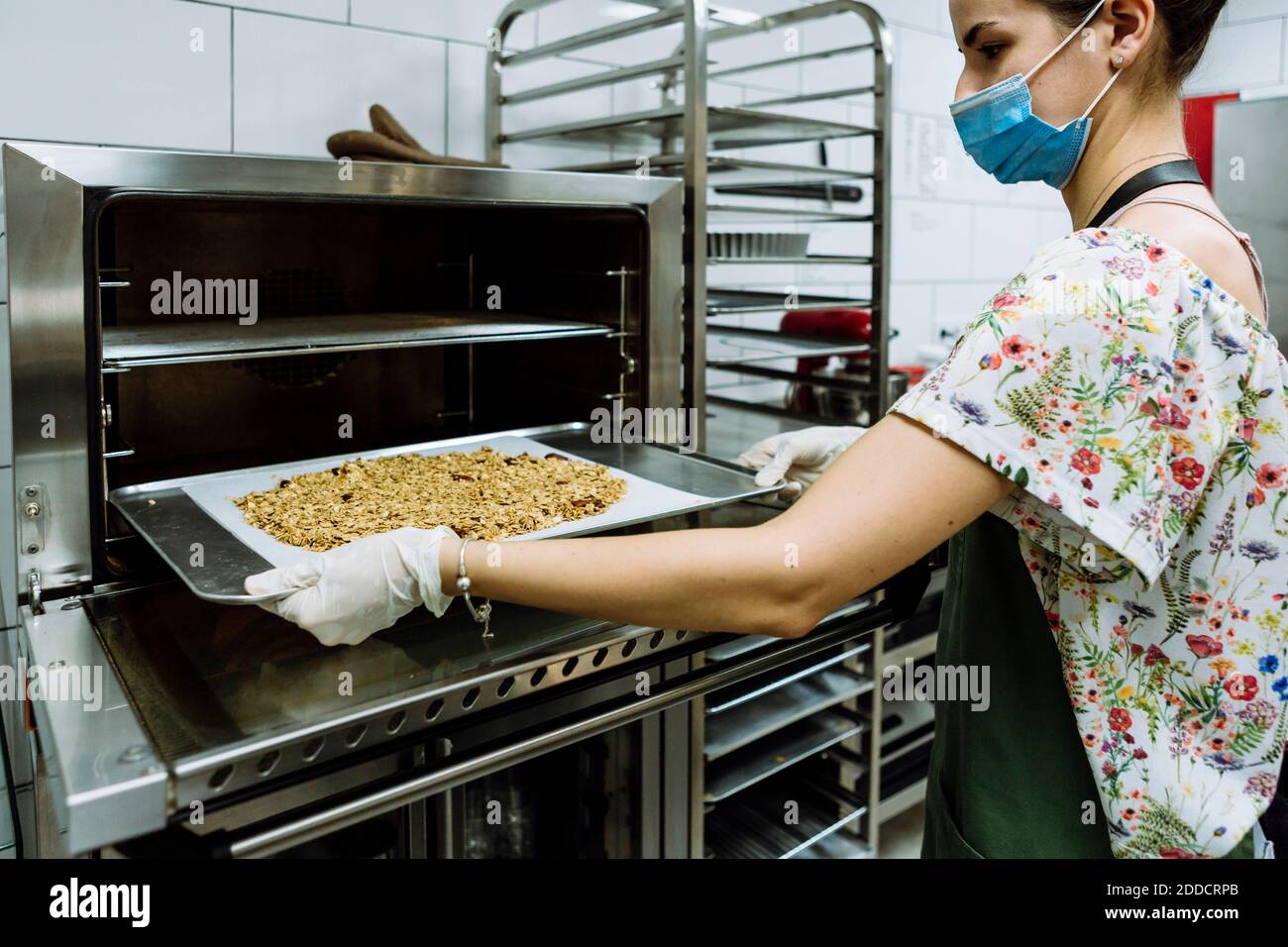 Junge weibliche Bäckerin Positionierung Blatt im Ofen in der Bäckerei Küche Während einer Pandemie Stockfoto