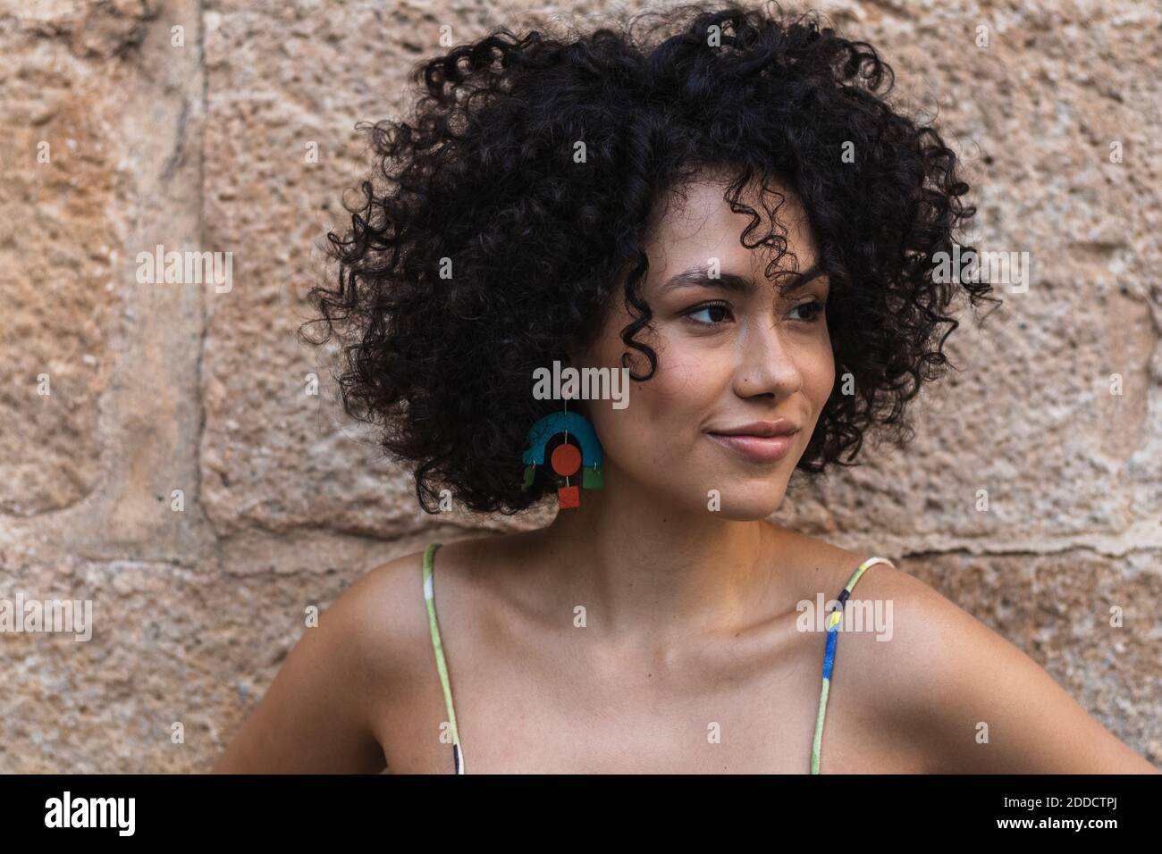 Lächelnde junge Frau schaut weg, während sie sich an die Steinmauer lehnt Stockfoto