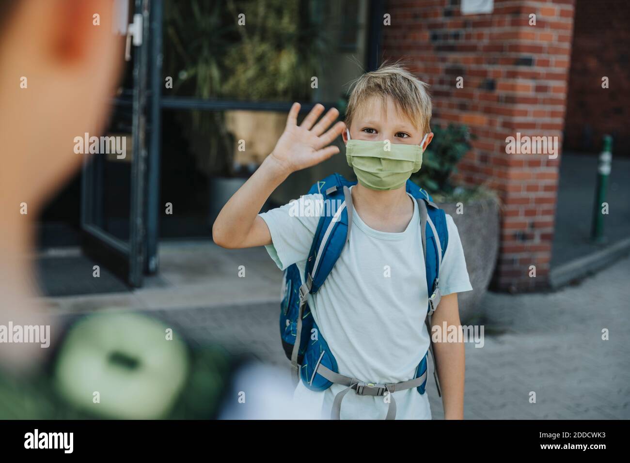 Kleiner Junge mit Schutzmaske, der seinen Bruder nach draußen schwenkt Schulgebäude Stockfoto