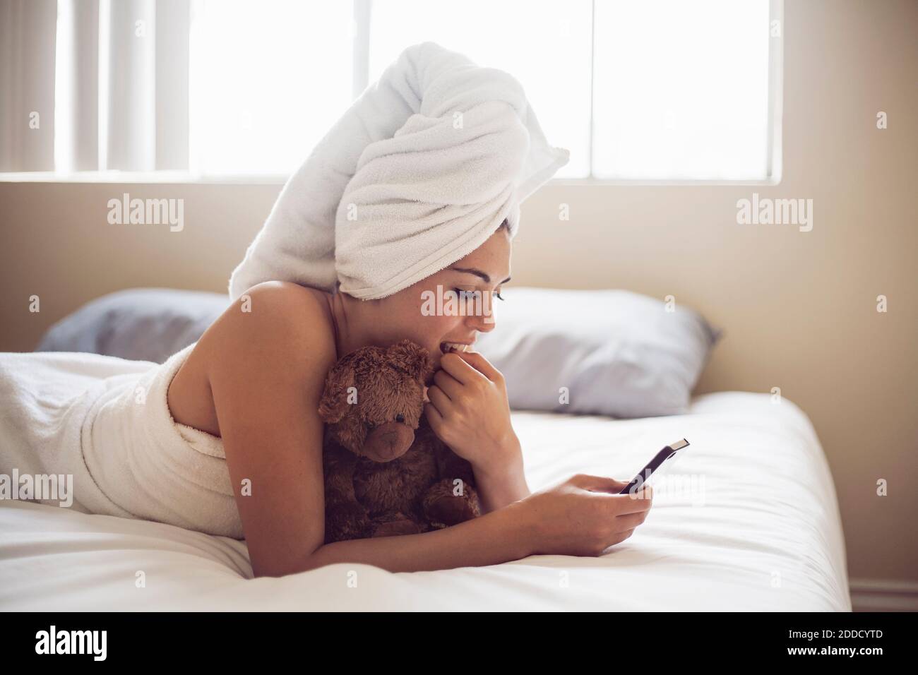 Junge Frau in Handtuch gewickelt mit Smartphone während liegen Auf dem Bett zu Hause Stockfoto