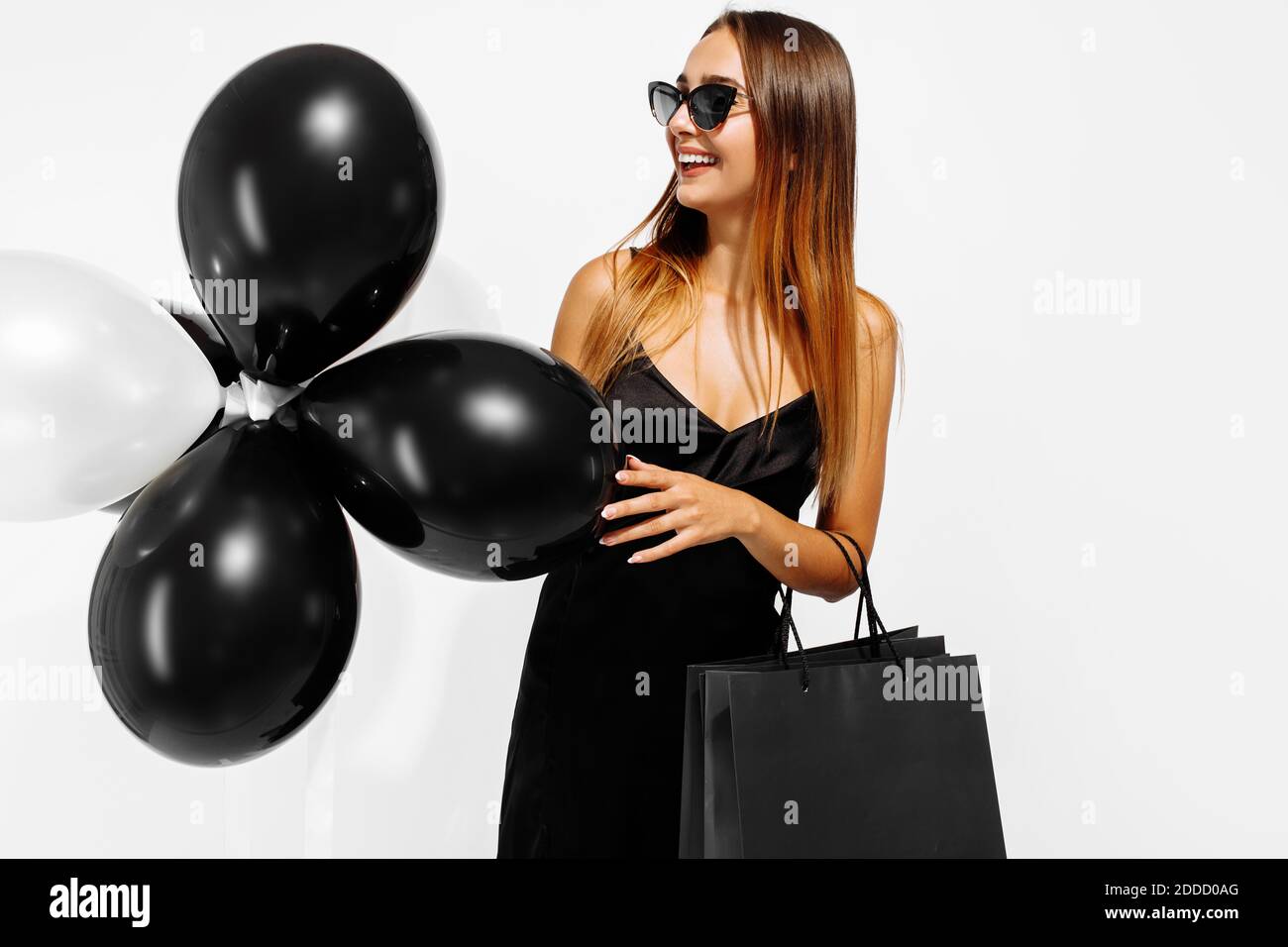 Black Friday Verkaufskonzept. Junge Frau in einem schwarzen Kleid, mit Luftballons und Einkaufstaschen, auf weißem Hintergrund Stockfoto